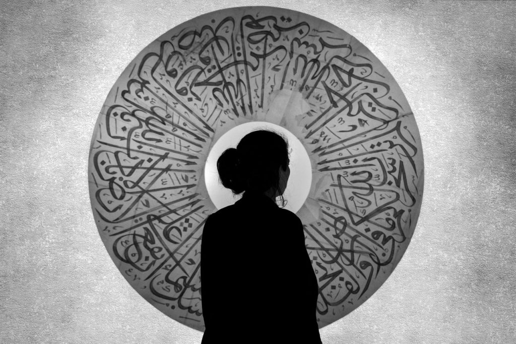 Silhouette of a woman standing before an ornate Arabic calligraphy artwork creating a striking monochrome effect.